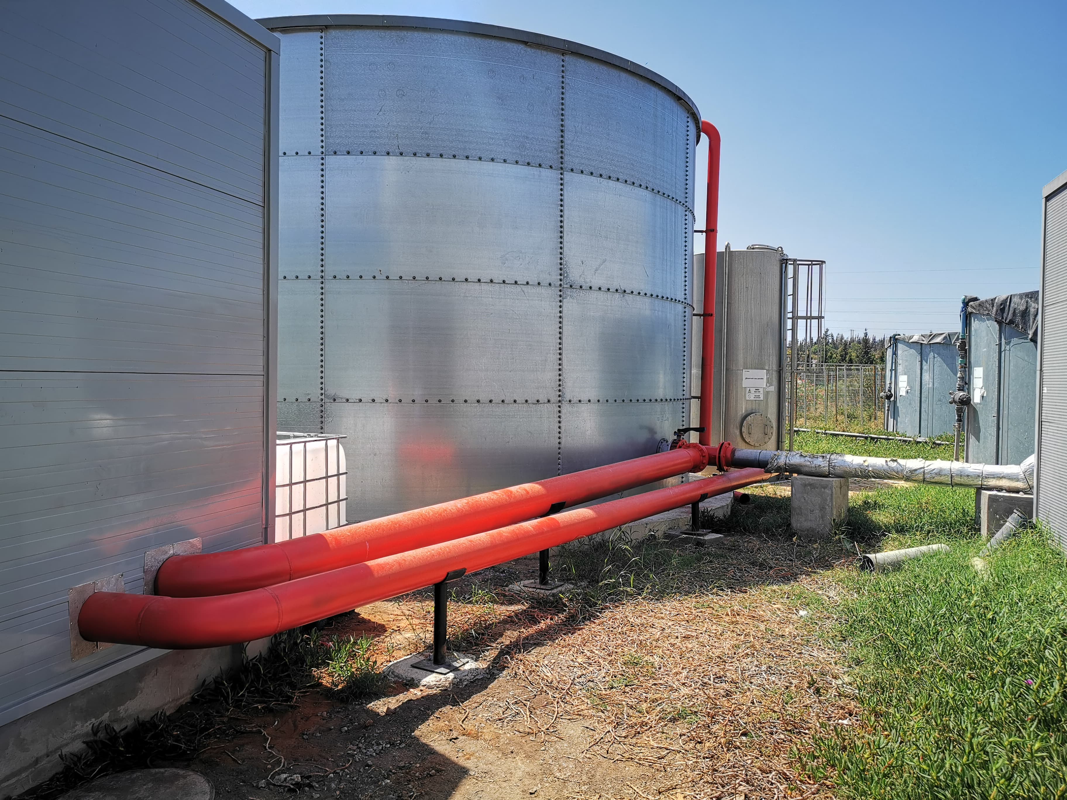 Grand réservoir métallique cylindrique avec deux tuyaux rouges horizontaux dans une installation industrielle extérieure.